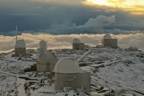 Obserwatoria astronomiczne na ośnieżonym szczycie, otoczone morzem chmur. W oddali widać złote światło zachodzącego słońca, a na pierwszym planie "la silla" - jedno z kopułowych budynków.